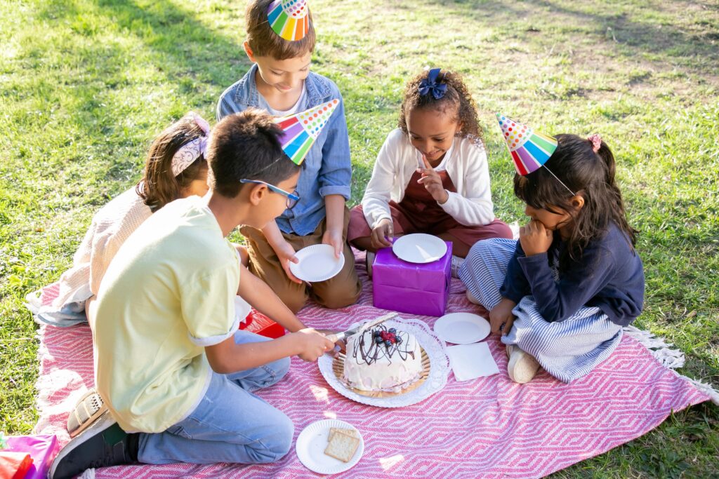 Five children sitting around a pink picnic blanket with rainbow coloured party hats
